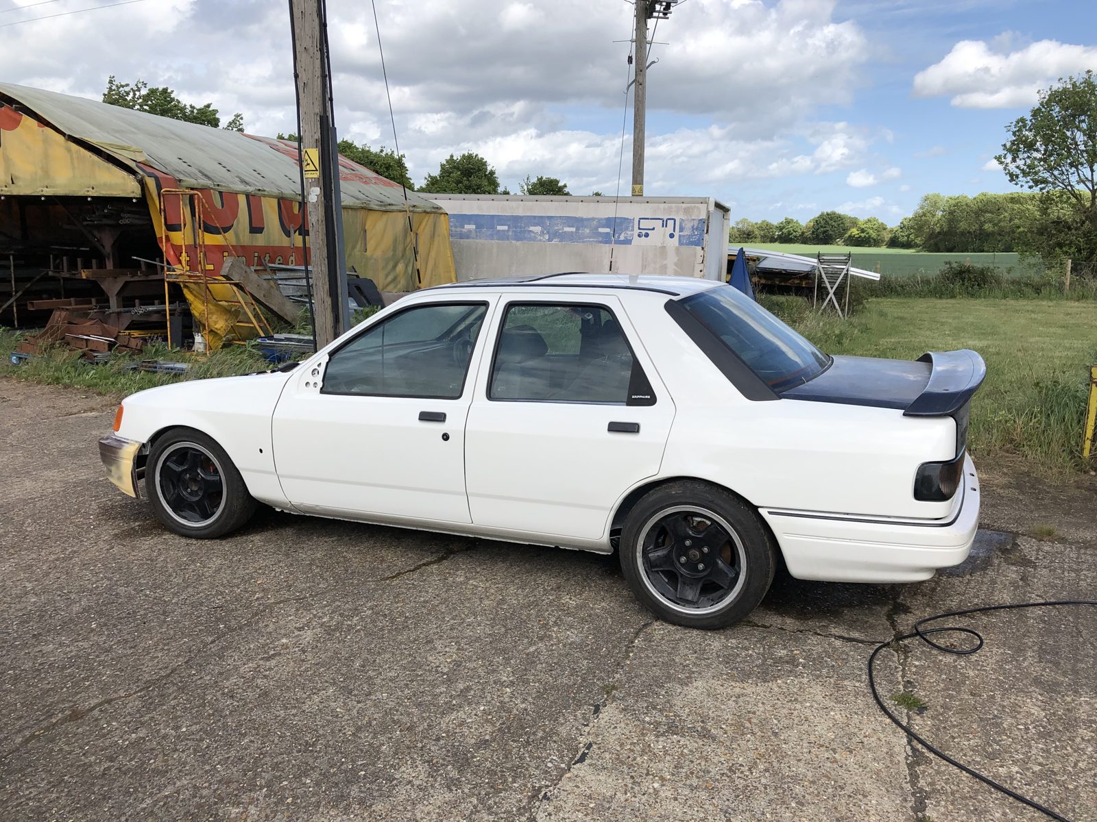 Side profile of the finished white Ford Sierra Sapphire Cosworth outdoors, with a black rear spoiler and black wheels